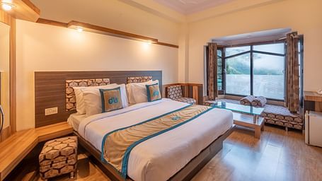 A hotel room with a large bed, a window overlooking a mountain view, and a seating area - DLS Meghavan Resort